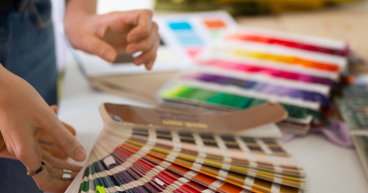 Designer holding a Pantone color guide fanned open next to printed color swatch books on a work surface