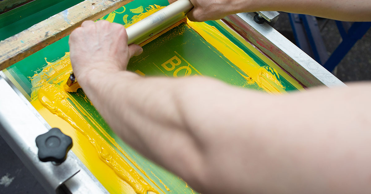 Close-up view of professional screen printing types demonstration showing hands using squeegee to apply vibrant yellow and green ink through mesh screen, displaying proper technique and pressure for quality screen printing results