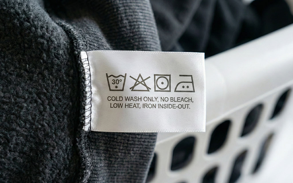 Close-up of hoodie care label showing laundry symbols and instructions: cold wash only, no bleach, low heat, iron inside-out to prevent print cracking